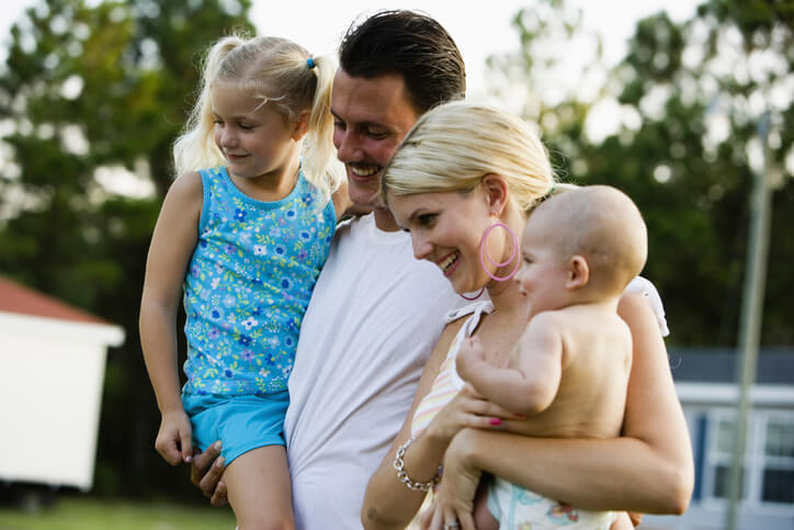 A mother and father hold their young children while standing outside their mobile home