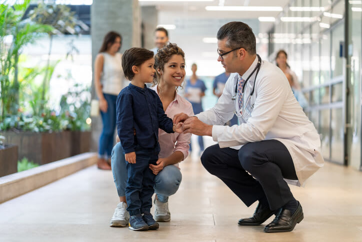 A doctor crouches to speak with young boy and his mother
