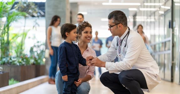 A doctor crouches to speak with young boy and his mother