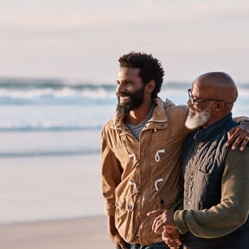 A father and adult son embrace and walk on the beach