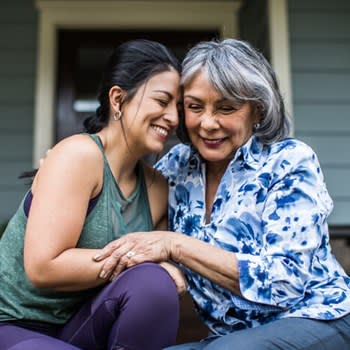 Woman and adult daughter embrace and smile while sitting on home front steps