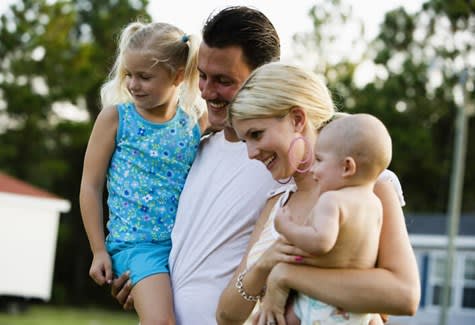 A mother and father hold their young children while standing outside their mobile home