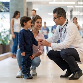 A doctor crouches to speak with young boy and his mother