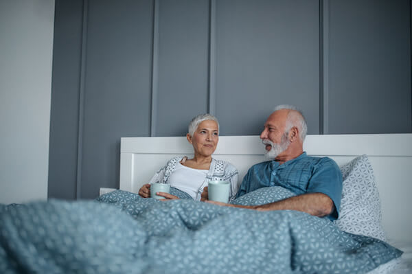A couple smiles and talks while drinking coffee in bed