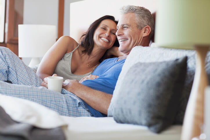 A couple smiles as they relax in bed with a cup of coffee
