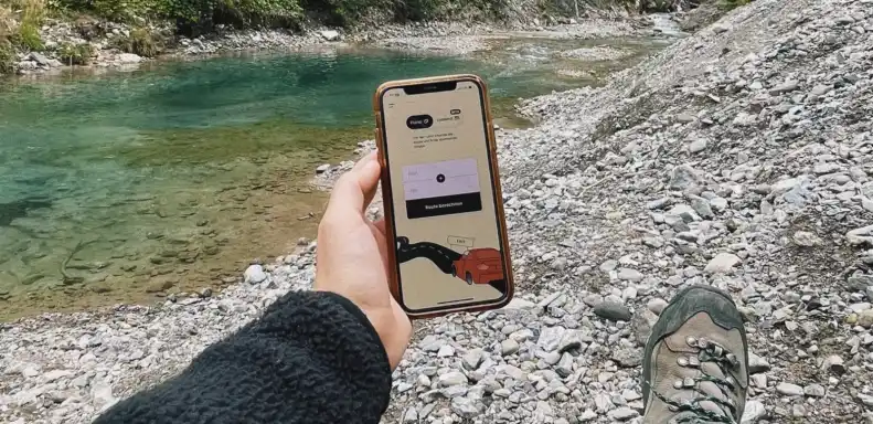holding a phone with outreached hand on the side of river with pebbles. On the phone is the by the ways app.