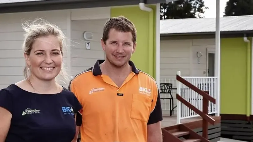 A smiling woman and man in BIG4 work shirts standing in front of a cabin with a lime-green wall at a holiday park in Launceston.