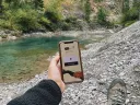 holding a phone with outreached hand on the side of river with pebbles. On the phone is the by the ways app.