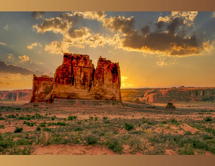 A landscape of the Courthouse Towers in Utah, featuring a large rugged rock formation with a reddish hue. illuminated by the light of the setting sun. The sky above is partly cloudy with the sun casting a golden glow on the clouds and surrounding terrain. The foreground consists of a sparse desert landscape with patched of green vegetation the scene evokes a sense of natural beauty and tranquility.