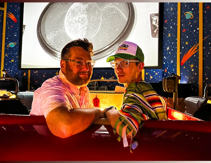 Matt and Nathan Kirouac-York, founders of The Gay Lane, sitting in an American diner booth themed like a red car. They are looking back at the camera with their arms resting on the back seat.