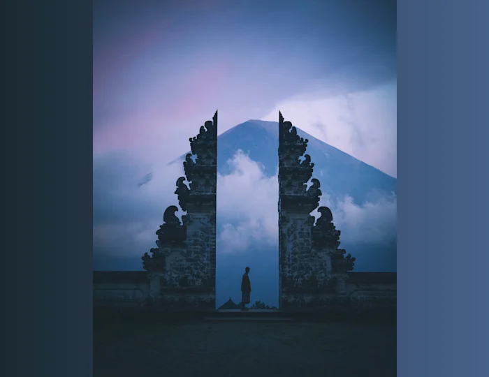 A person standing near an ancient stone structure with a mountain and mist in the background with purple sky