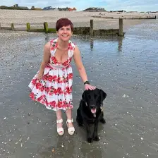 Sassy is a petite white woman, wearing a sleeveless dress with a floral pattern of predominantly red roses with green leaves on a white background. She has short plum hair and is smiling warmly as she crouches on East Wittering Beach with her hand atop her guide dog Ida's head. Ida is a black Labrador retriever cross and her pink tongue is poking out making her look very happy indeed.