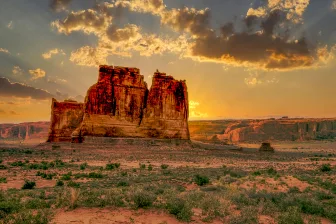 A landscape of the Courthouse Towers in Utah, featuring a large rugged rock formation with a reddish hue. illuminated by the light of the setting sun. The sky above is partly cloudy with the sun casting a golden glow on the clouds and surrounding terrain. The foreground consists of a sparse desert landscape with patched of green vegetation the scene evokes a sense of natural beauty and tranquility.