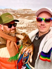 Matt and Nathan holding their Dachshund dog outside with the backdrop of a national park with layered smooth rocky hills behind them.