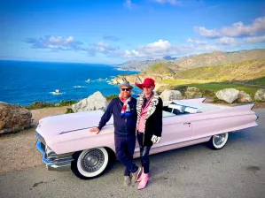 Matt and Nathan standing in front of a 1950s pink convertible car with a scenic view of the blue ocean and green coastline behind them.