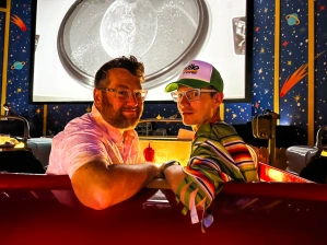 Matt and Nathan Kirouac-York, founders of The Gay Lane, sitting in an American diner booth themed like a red car. They are looking back at the camera with their arms resting on the back seat.