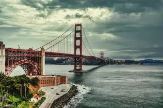 A photo of the Golden Gate Bridge under cloudy skies. Sunrays are popping through the clouds. On the left of the frame is the structures that the road sits on. Across the cam waters of the bay the Marin Headlands can be seen.