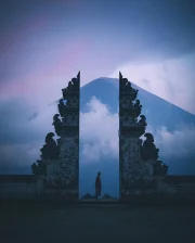 A person standing near an ancient stone structure with a mountain and mist in the background with purple sky