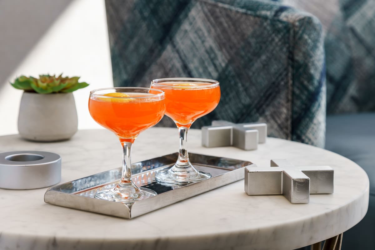 Two orange color drinks on a silver tray with big X and O signs and a vase placed on the table.