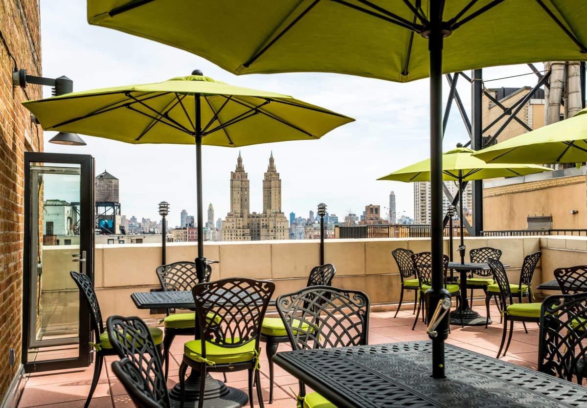 A rooftop patio with yellow umbrellas and black metal tables and chairs, overlooking the New York City skyline.