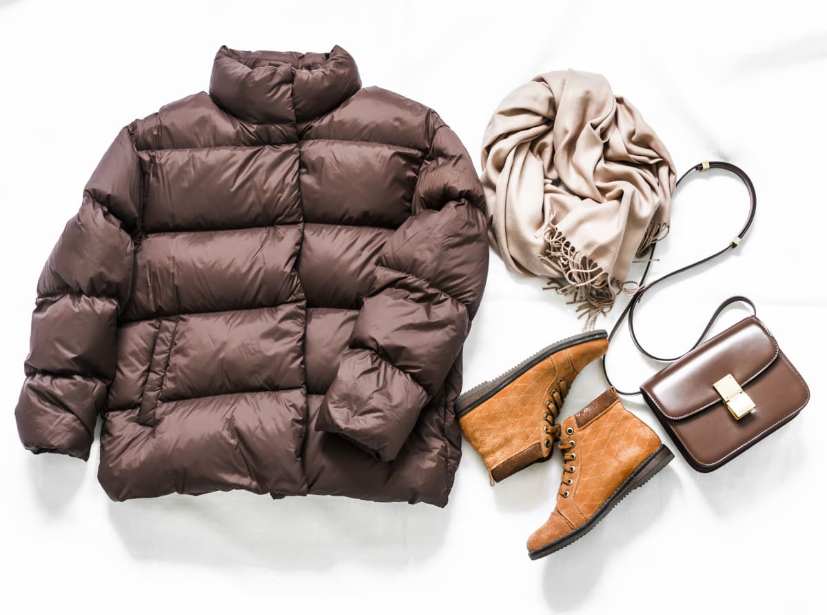 stock photo of brown down puffer jacket, orange ankle boots, a cream colored scarf and a small brown satchel bag on a white background. 