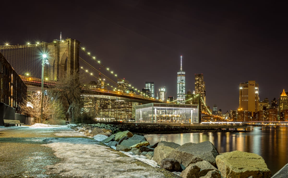 Snow covered sidewalk leading towards Brooklyn Bridge at night time. Bridge lit up with the New York city lights in the background. 