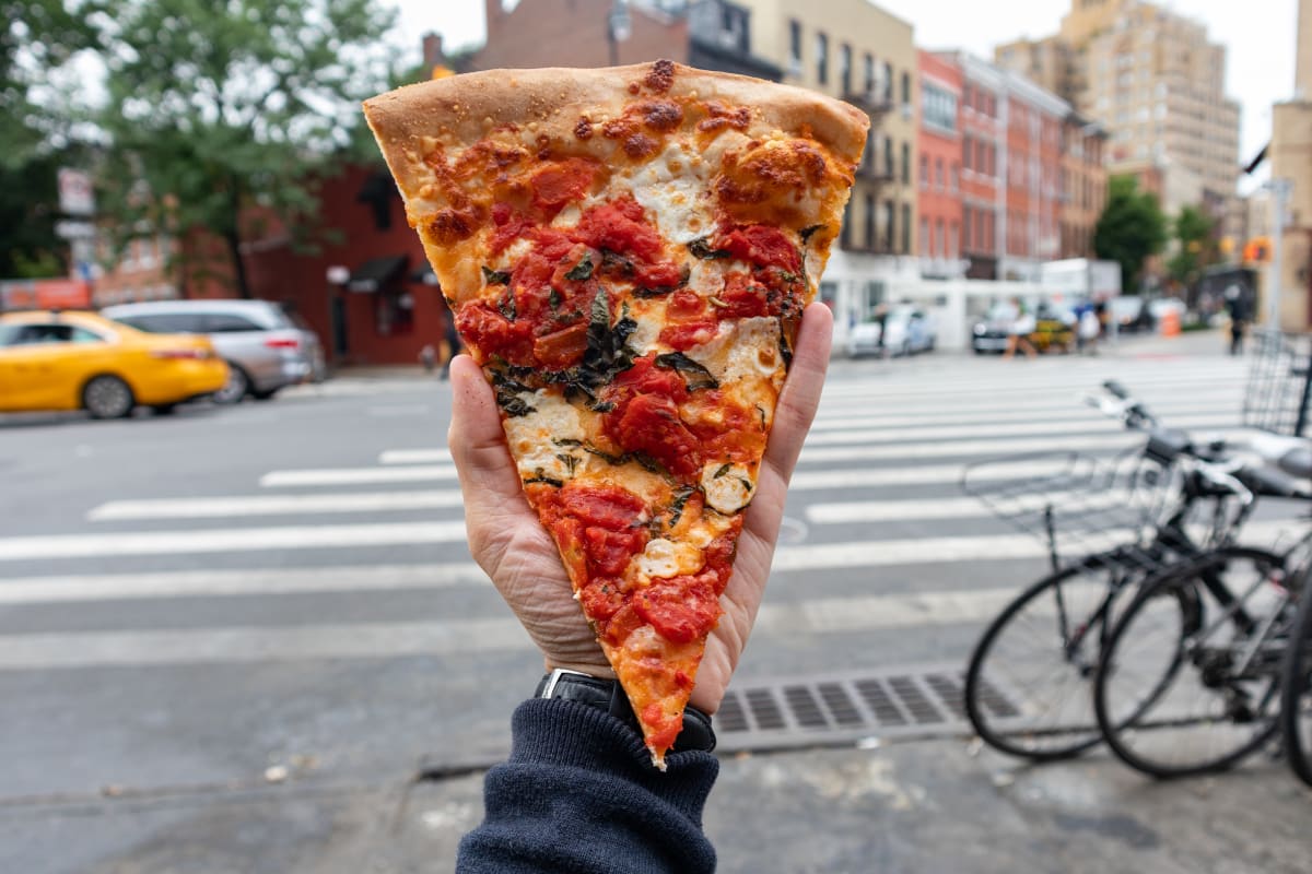 A slice of New York pizza held up in front of a NYC intersection. 