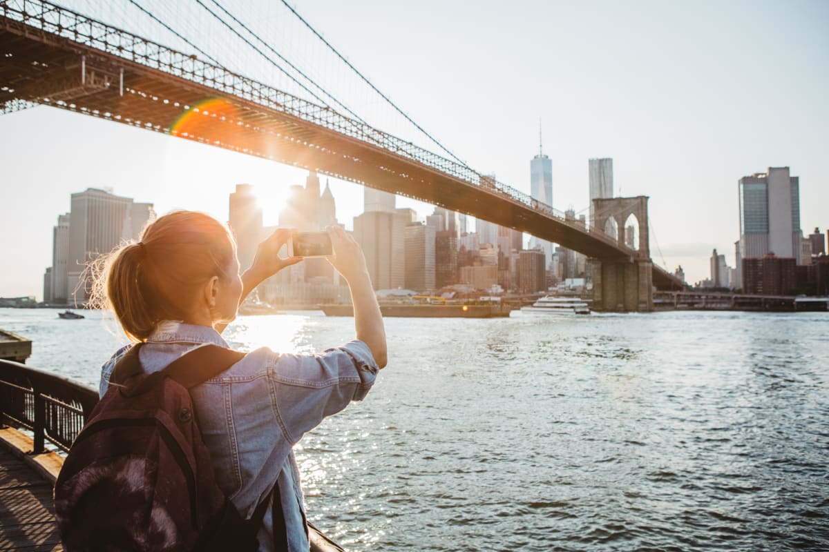 Female tourist with backpack takes photo of Manhattan skyline from the river bank under the Brooklyn Bridge  