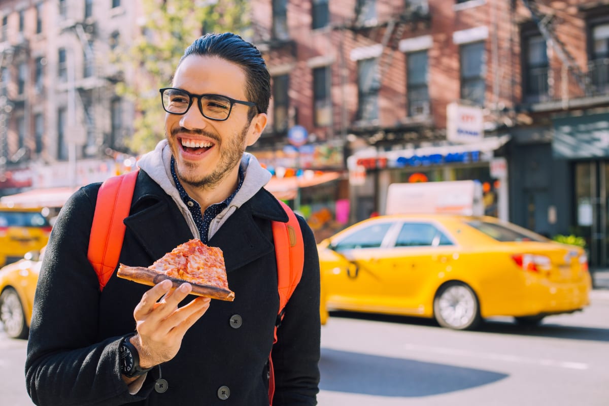 Young man with glasses and beard smiles as he eats a slice of new york style pizza, on a new york street in front of yellow taxi cab