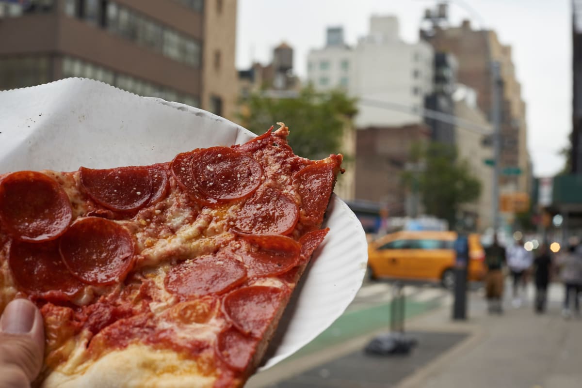 slice of new york style peperoni pizza, with a slice taken out on a paper plate with a new york taxi and street in the background. 