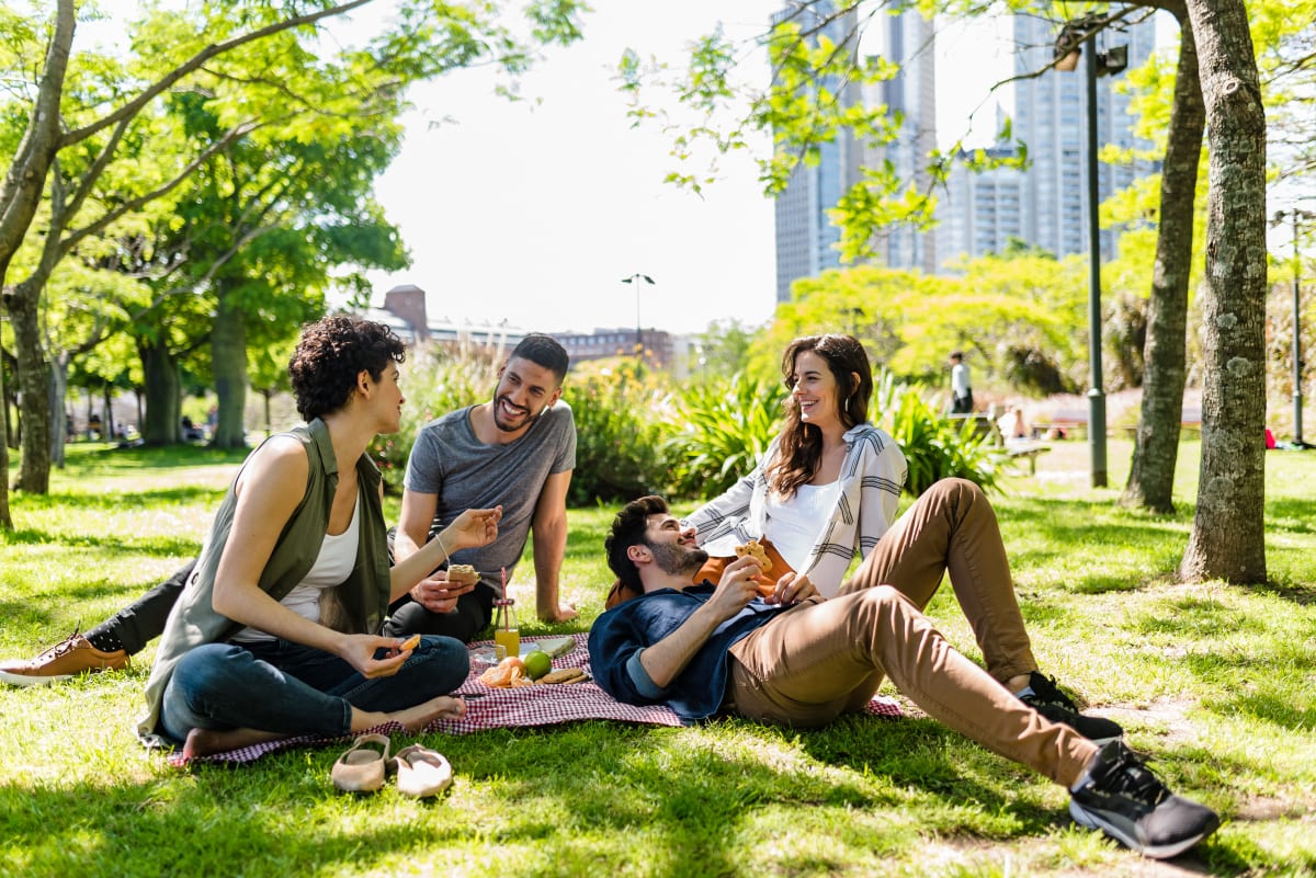 Friends sit on a picnic blanket in central park laughing, trees and buildings in the background