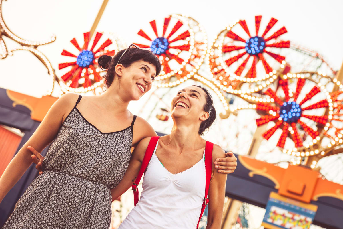 Two women walk arm in arm smiling with a large red, white and blue fairground attraction in the background. 