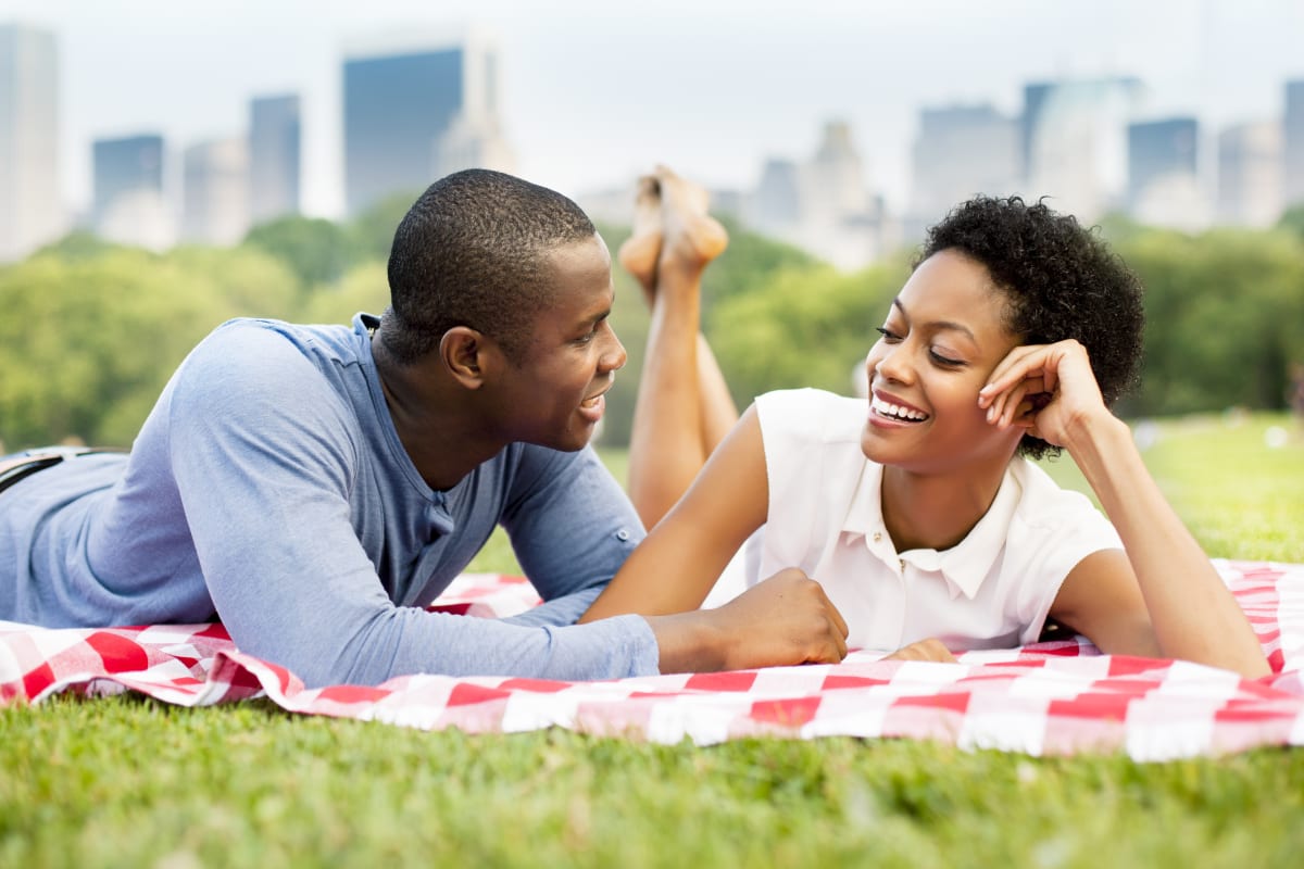 Man and woman smile as they look at each other, while laying side by side on a red and white picnic blanket with the new york city skyline out of focus in the background