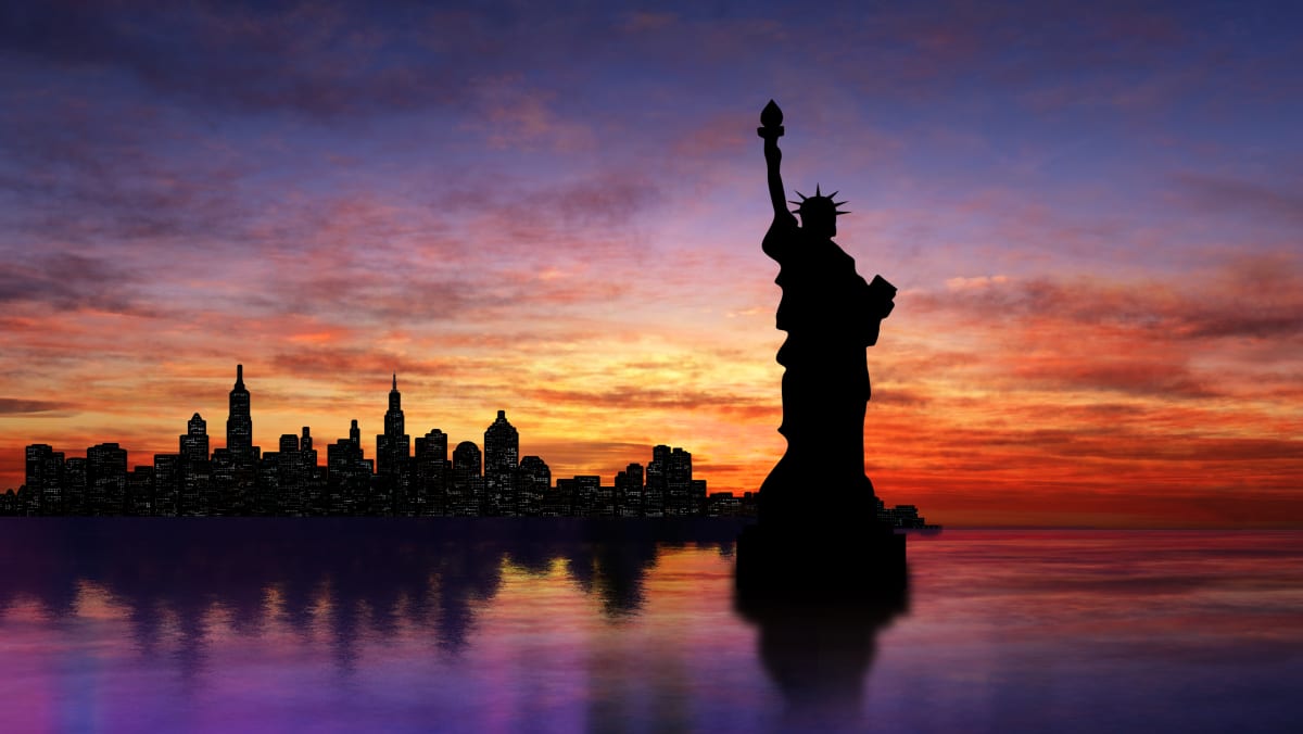 Silhouette of the Statue of Liberty, with a sunset and the new york skyline in shadow in the background 