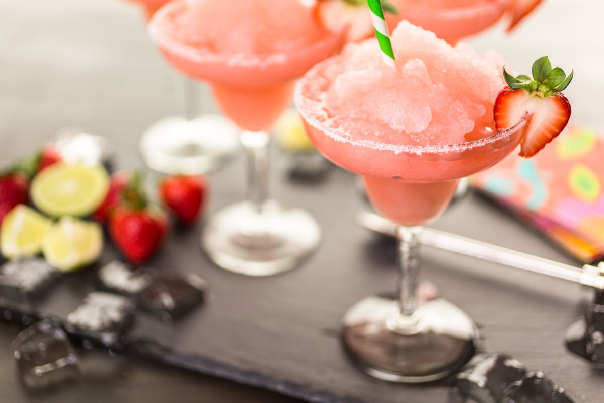 Two pink frozen cocktails in margarita glasses with a salted rim, green and white striped straws and a strawberry garnish, fruits are out of focus in the background of the image