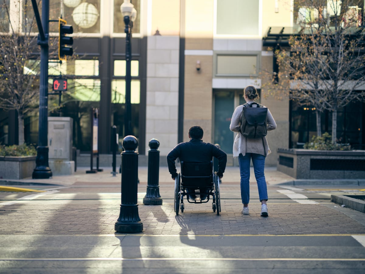 man using a wheelchair next to a woman walking across a street with buildings in the background