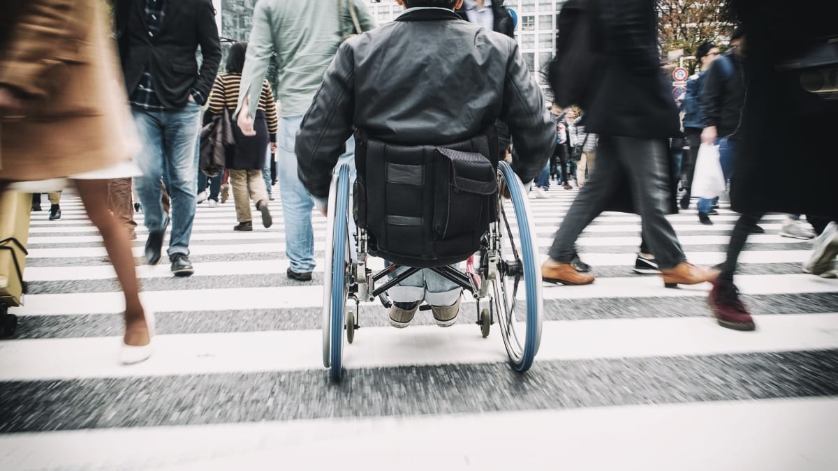 man is wheelchair crosses cross walk between lots of people walking and rushing across the same crossing 