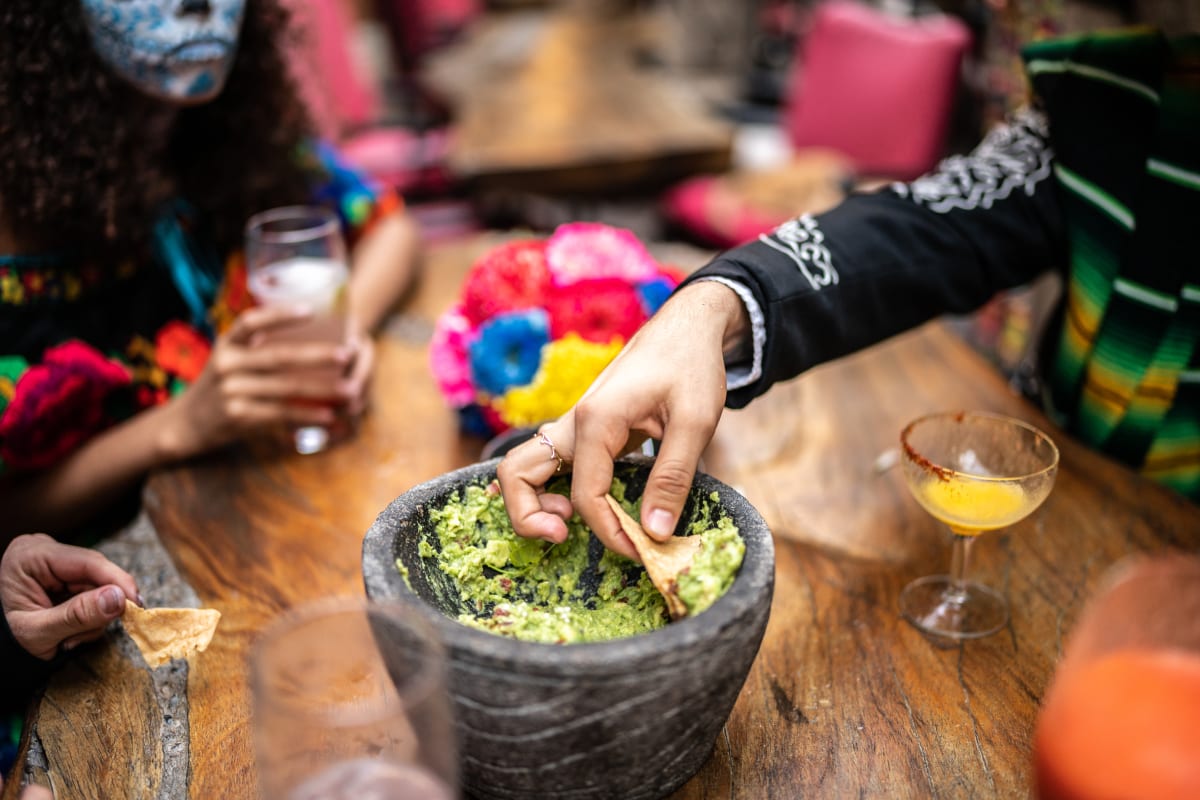 Bowl of guacamole on a table, with hand dipping tortilla chip inside, day of the dead flowers and a drink are also on the table