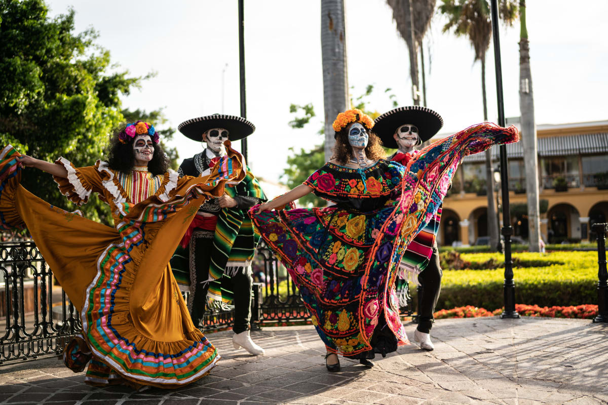 Men and women dressed in traditional day of the dead clothing, with traditional headwear, dancing under palm trees