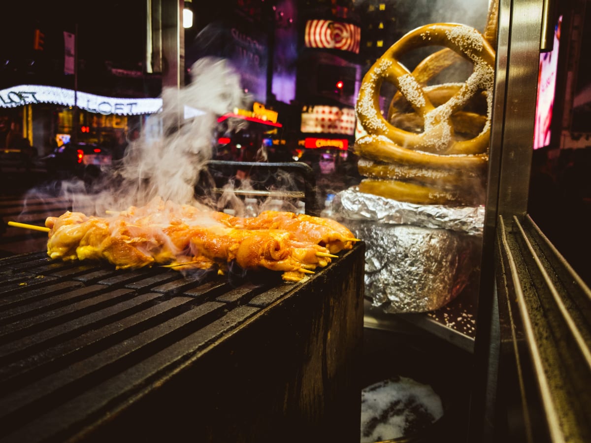 food cooking on grill with giant pretzels and the lights of new york city in the background