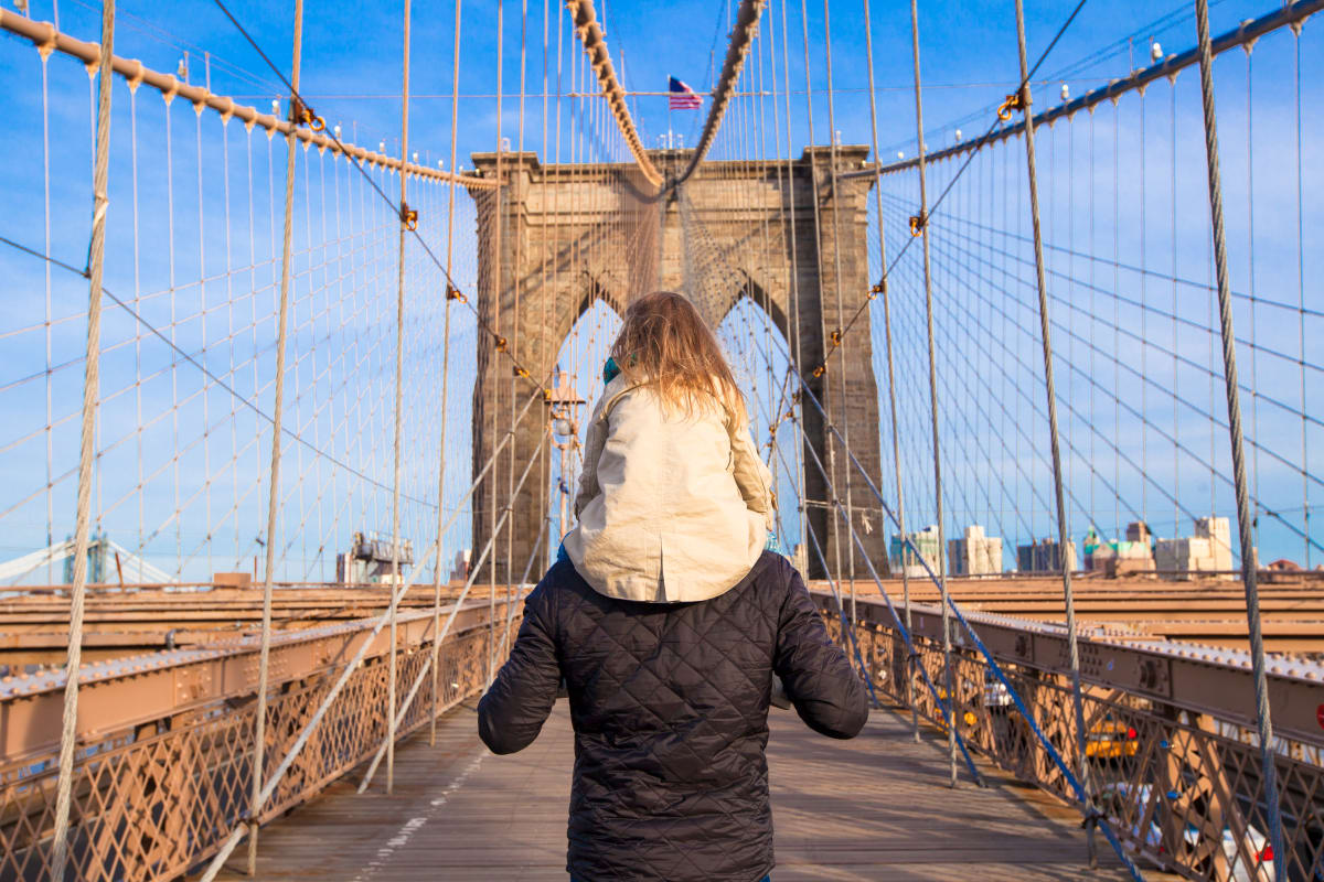 Young girl sits on dads shoulders as they cross Brooklyn Bridge