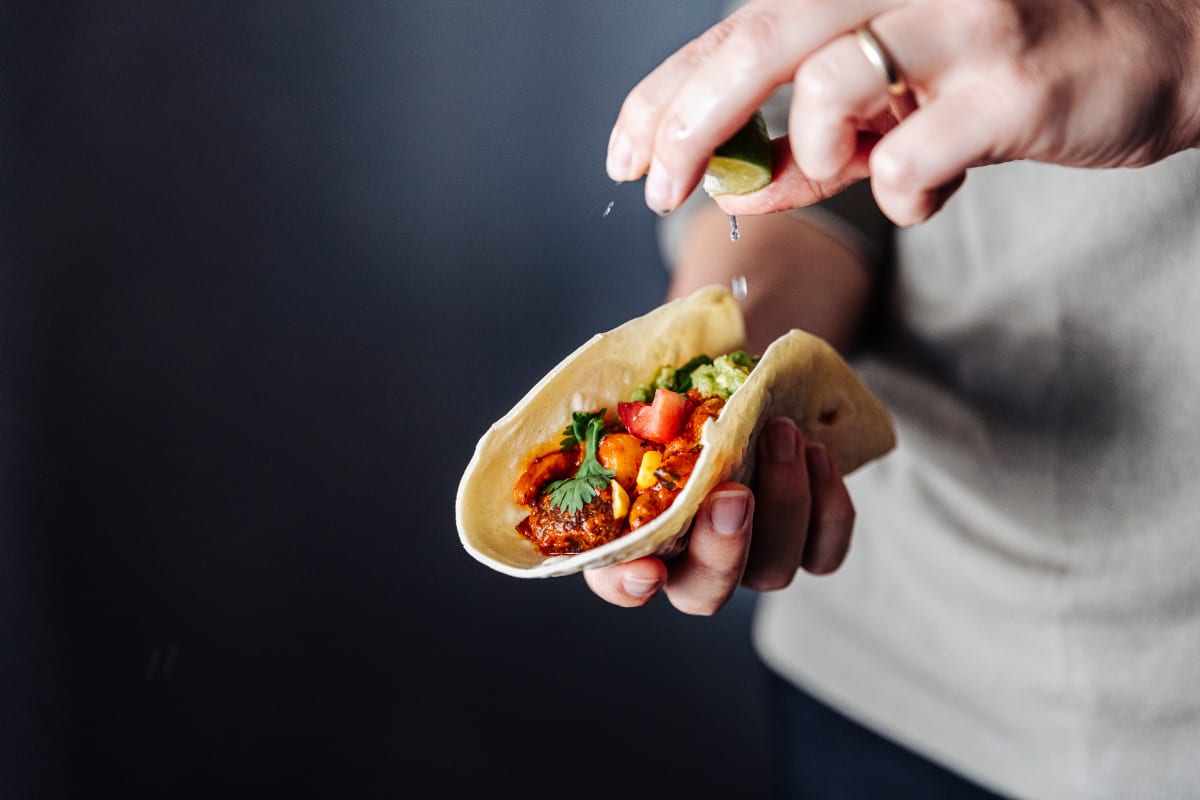 Vegan taco in someone's hand, vegetables and coriander inside the taco with a second hand, wearing a silver ring, squeezing lime into the taco.  