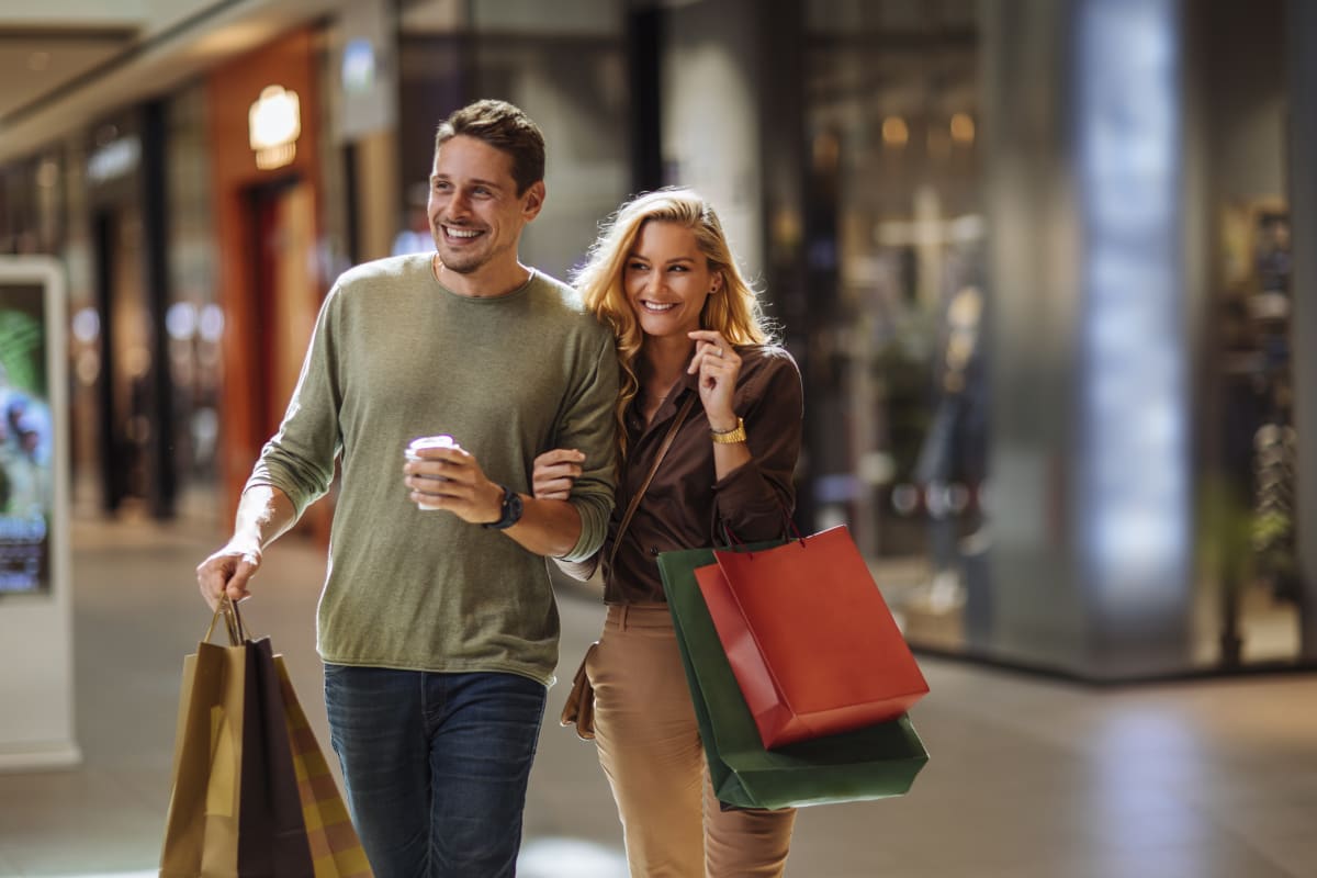 Couple linked arm in arm, with lots of shopping bags. Mall stores blurred in the background. 