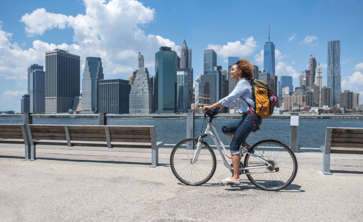 Woman cycles in the sunshine, beside benches on the Hudson river with the new york sky line in the background 