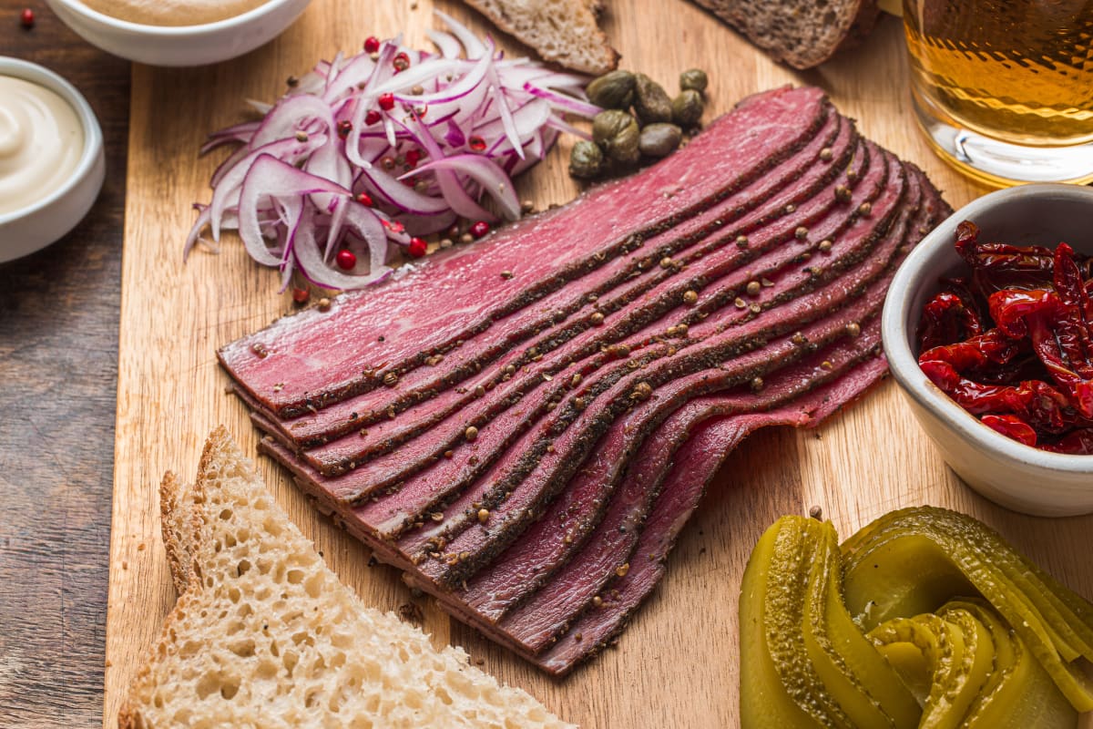 Sliced Pastrami meat on a chopping board with sliced red onion, capers and peppercorns, alongside sliced bread, pickles, a red vegetable and a variety of sauces in the background