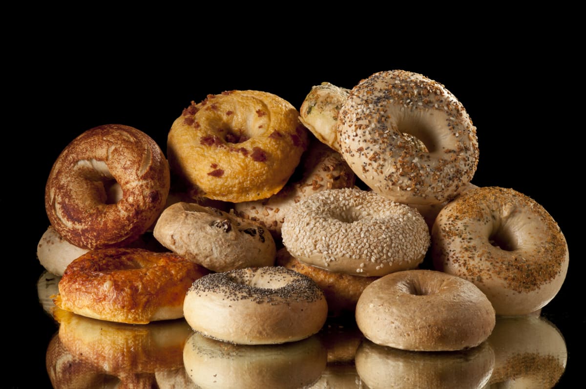 A variety of bagels piled onto a black mirrored surface, with black background. Some bagels have poppyseeds, others sesame and others cheese. 