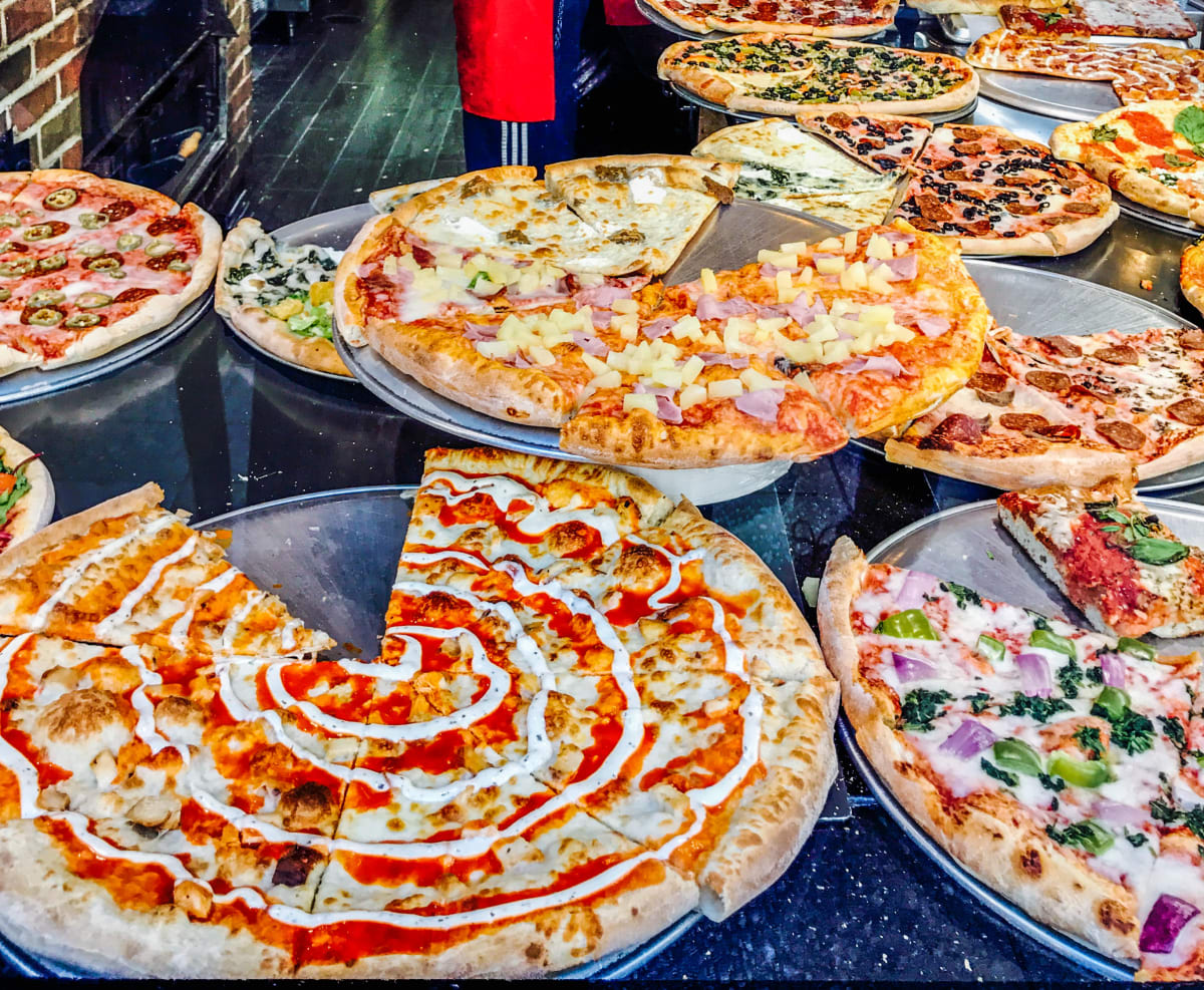 New York Style Pizza, sold by the slice, on a back granite counter top, with about 15 pizza, all with different toppings. 