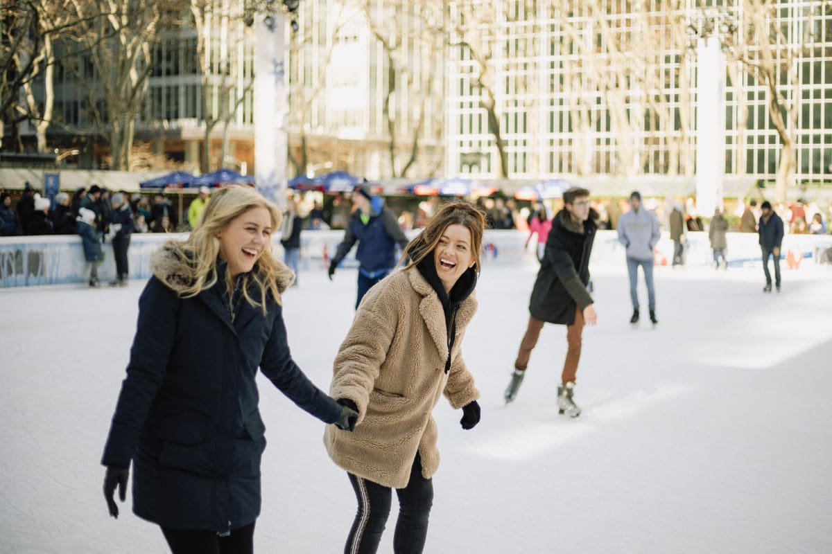 Two women in coats iceskate at Wollman Rink, smiling to each other 