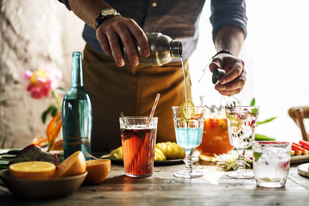 Man makes a cocktail using a metal shaker, various glasses are filled with different coloured drinks, there are other cocktail ingredients on the table 