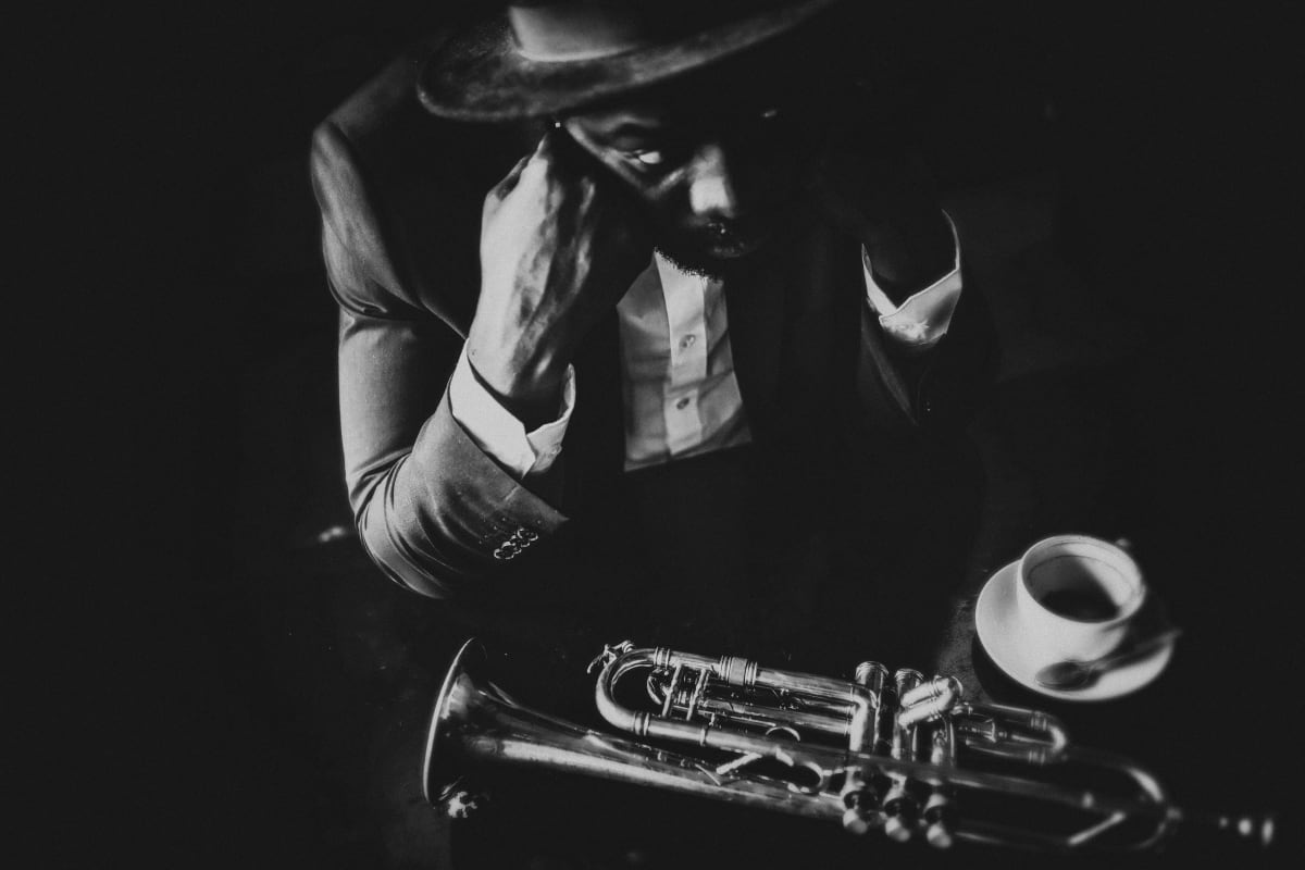 Black and white photo of Saz player with a coffee and sax on the table, no background visible, player is wearing suit, bow tie and hat 